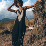 A person dressed in a long black Boho Dress by AYA Sacred Wear, featuring fringed tribal details, is posing outdoors while leaning against a large rock. The backdrop showcases dry grass, rocky terrain, and a partly cloudy blue sky.
