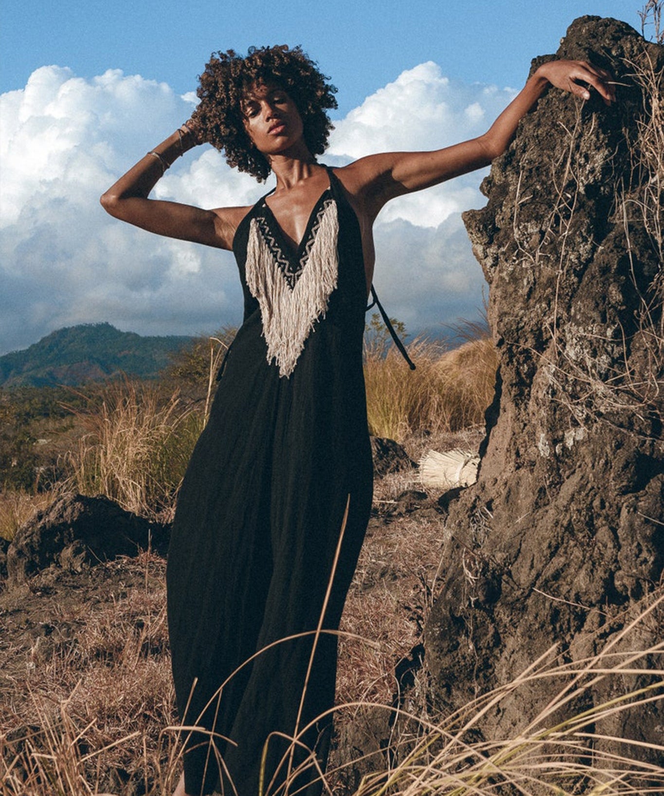 A person dressed in a long black Boho Dress by AYA Sacred Wear, featuring fringed tribal details, is posing outdoors while leaning against a large rock. The backdrop showcases dry grass, rocky terrain, and a partly cloudy blue sky.