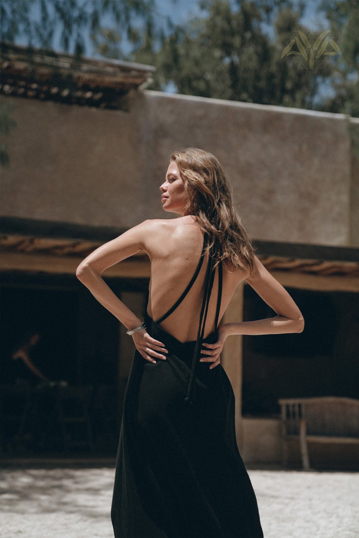 A woman stands outdoors in front of a rustic building, wearing a backless black dress from the AYA Sacred Wear collection. Her long hair flows down her back as she looks to the side. The scene is sunlit with greenery in the background and soft, natural tones.