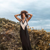 A person with curly hair poses confidently outdoors, wearing an AYA Sacred Wear black Boho Dress, featuring a fringed front detail. They stand on a stone surface, surrounded by trees under an overcast sky.