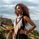 A woman with curly hair stands confidently outdoors near the ocean, beautifully dressed in a fringed top, adorned with necklaces and bracelets. She's wearing an organic bohemian dress from AYA Sacred Wear's collection, perfect for any event. The background showcases rocky terrain, lush greenery, and a cloudy sky.