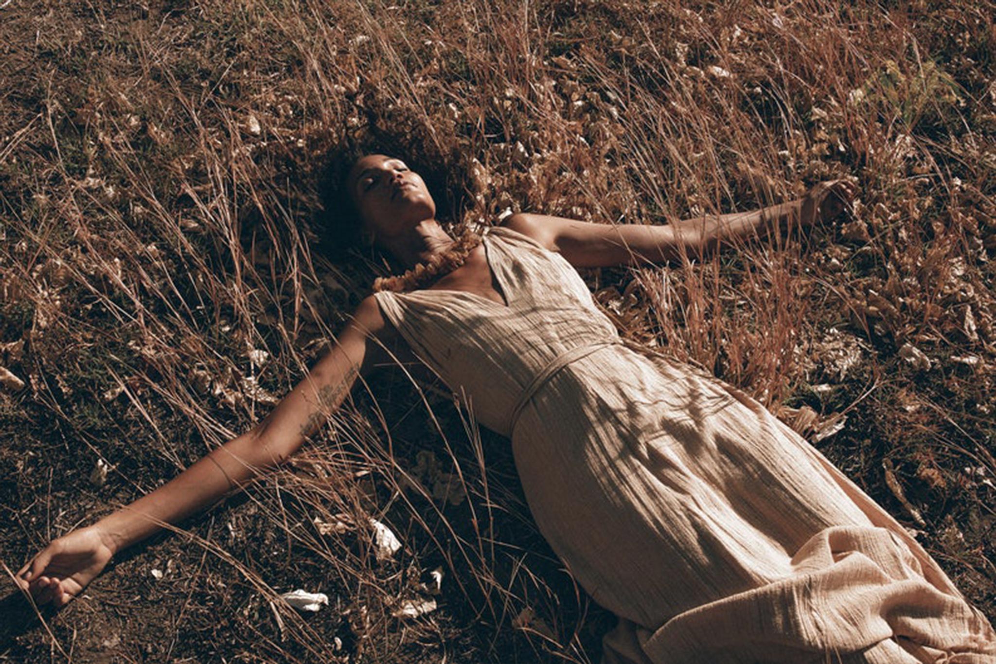 A person in an AYA Sacred Wear Boho Dress for Woman — a long maxi bohemian style in beige sandal organic fabric — lies peacefully on the dry, grassy ground with arms outstretched, surrounded by brown plants. The warm lighting adds a serene, autumnal atmosphere.