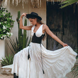 A woman in a wide-brimmed hat poses gracefully outdoors near a thatched-roof structure and cacti, wearing the Off-White Maxi Adjustable Bohemian Dress by AYA Sacred Wear. She pairs it with black boots and holds the dress's hem, showcasing its movement and design.
