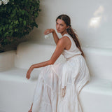 A woman dressed in an off-white Boho Dress by AYA Sacred Wear sits on a white bench, holding a small flower. She gazes thoughtfully to the side, surrounded by lush green plants in a serene setting.