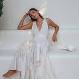 An elegant woman, wearing the Off-White Maxi Adjustable Dress from AYA Sacred Wear's Bohemian collection, sits gracefully on a minimalist white bench. Her head rests thoughtfully on her hand as she gazes contemplatively into the distance. Beside her lies a large white seashell, while green foliage nearby enhances the scene's tranquil charm and organic cotton elegance.