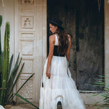 A woman in an off-white maxi Boho Dress by AYA Sacred Wear, complemented by a black hat, stands in front of a rustic wooden door amidst tall green cacti and textured walls under a thatched roof.