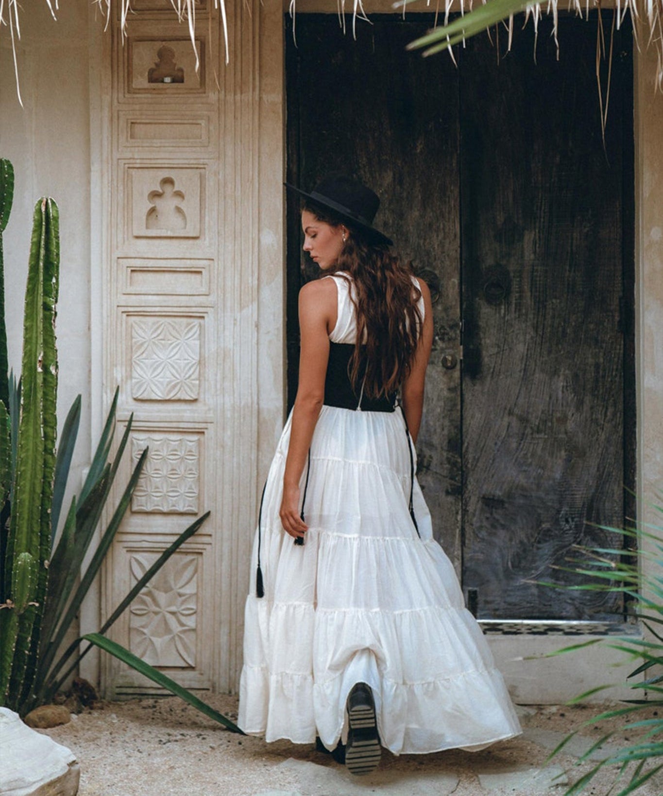 A woman in an off-white maxi Boho Dress by AYA Sacred Wear, complemented by a black hat, stands in front of a rustic wooden door amidst tall green cacti and textured walls under a thatched roof.