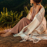 A woman sits on the grass, wrapped in an AYA Sacred Wear Boho Fringe Shawl • Cotton Scarf • Organic Wedding Shawl, gazing into the distance with lush greenery behind her. She wears a sleeveless, flowing dress and appears relaxed amidst the natural setting.