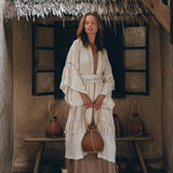 In a rustic setting with a thatched roof and wooden bench, an individual is seen wearing the Boho Kimono Cardigan by AYA Sacred Wear. This layered, textured robe harmonizes traditional and contemporary elements as they hold a decorative jug and sport white boots.