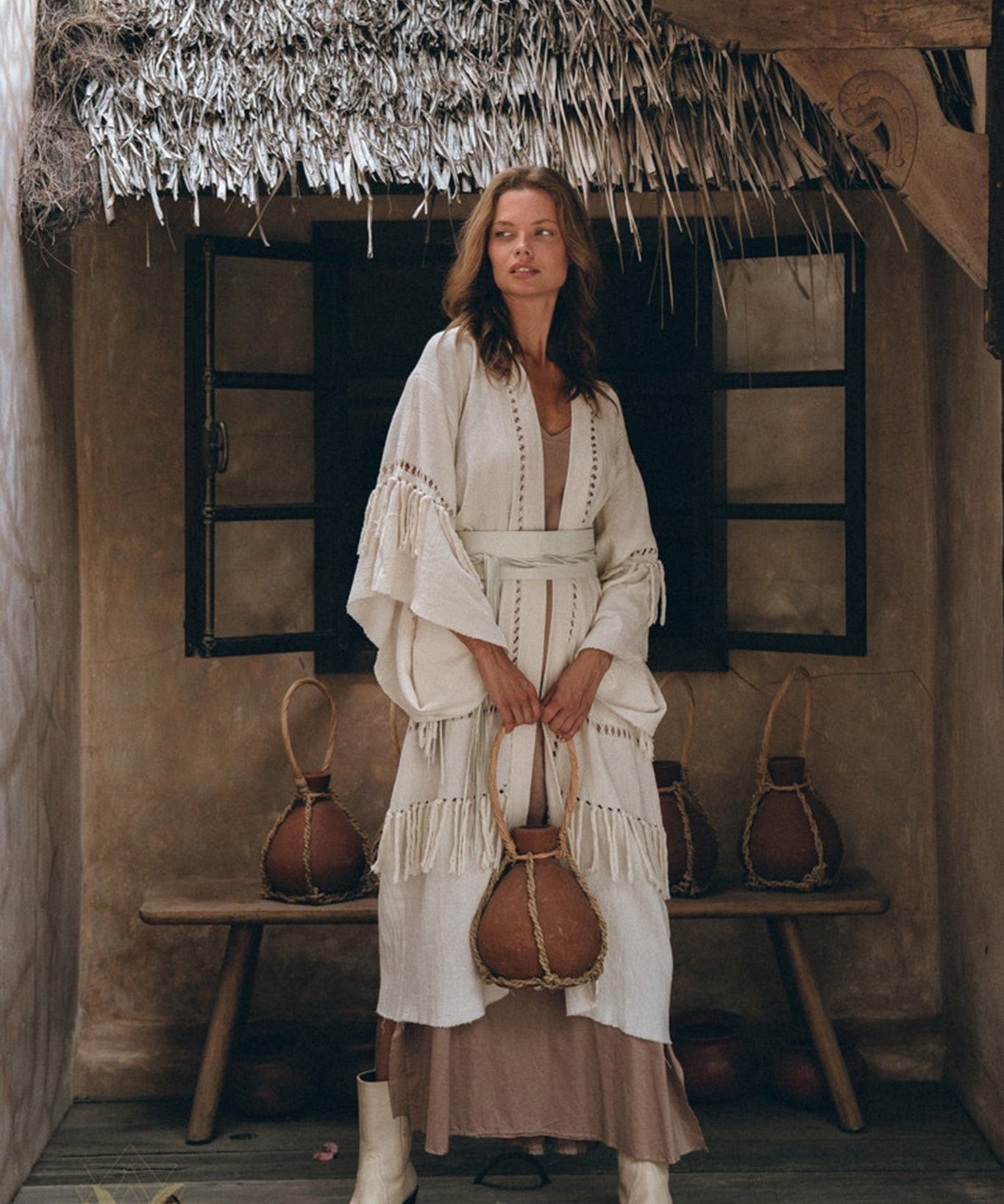 In a rustic setting with a thatched roof and wooden bench, an individual is seen wearing the Boho Kimono Cardigan by AYA Sacred Wear. This layered, textured robe harmonizes traditional and contemporary elements as they hold a decorative jug and sport white boots.