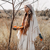 A woman wearing a handloom kimono from AYA Sacred Wear's Boho Kimono Cardigan collection stands in a grassy field, holding a tray of oranges. She has long hair and large earrings, gazing thoughtfully to the side near a leafless tree under the bright, clear sky.