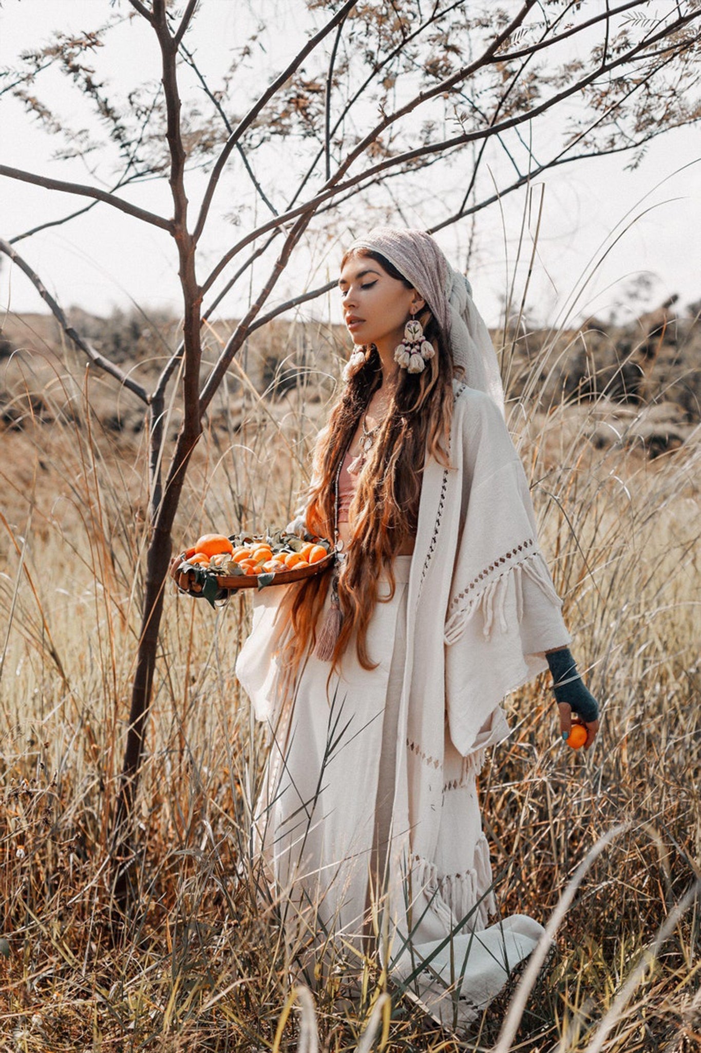 A woman wearing a handloom kimono from AYA Sacred Wear's Boho Kimono Cardigan collection stands in a grassy field, holding a tray of oranges. She has long hair and large earrings, gazing thoughtfully to the side near a leafless tree under the bright, clear sky.