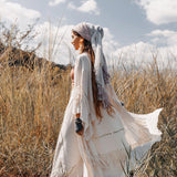 A woman in a flowing white dress and the Boho Kimono Cardigan by AYA Sacred Wear walks through tall grass in a sunny, open field. The sky is blue with scattered clouds, creating a serene, bohemian atmosphere.