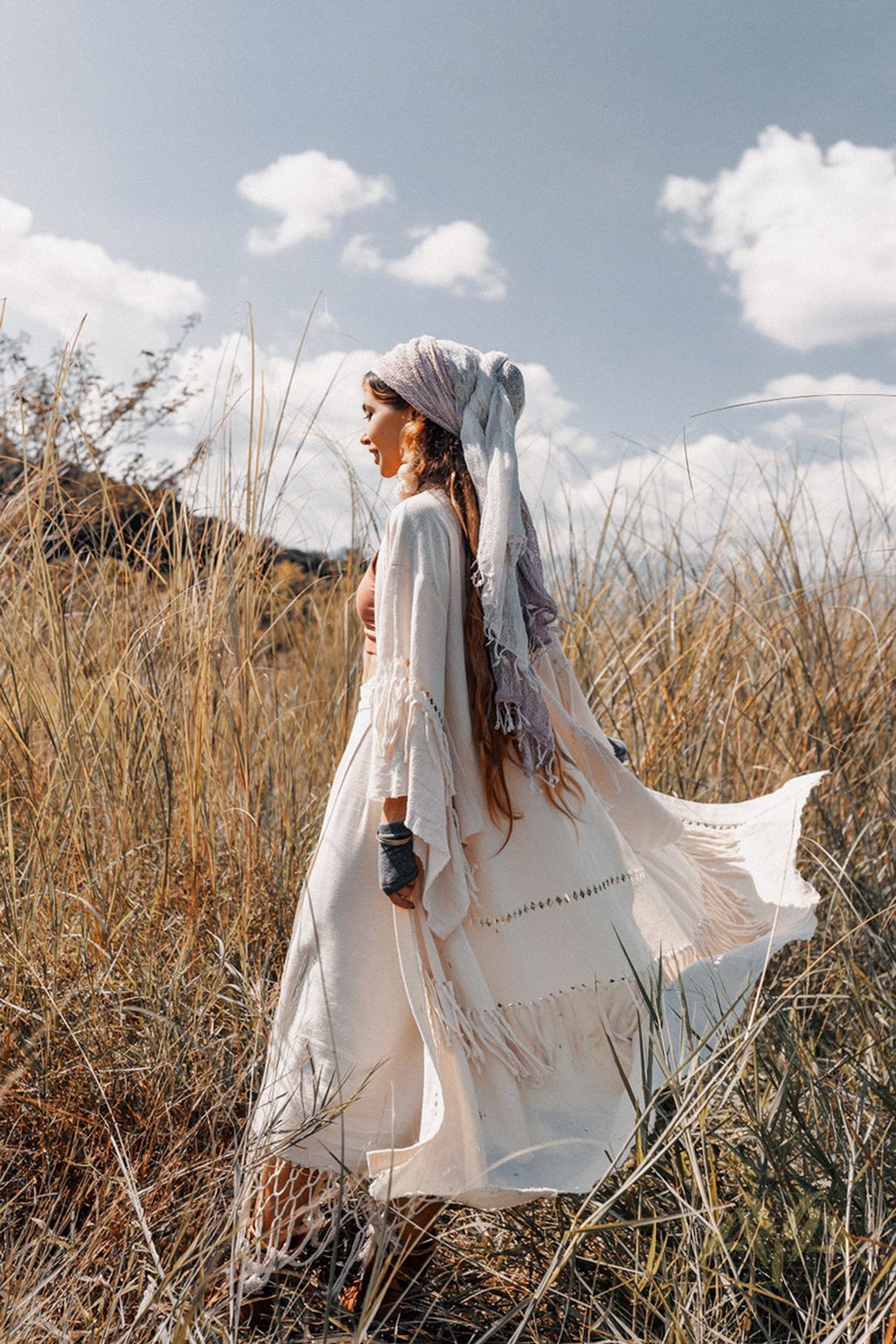 A woman in a flowing white dress and the Boho Kimono Cardigan by AYA Sacred Wear walks through tall grass in a sunny, open field. The sky is blue with scattered clouds, creating a serene, bohemian atmosphere.