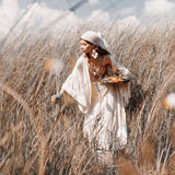 A woman dressed in a white dress and shawl gracefully walks through a field of tall grass, wearing the AYA Sacred Wear's Boho Kimono Cardigan complemented by a stylish headscarf. She gazes downward while holding a bouquet of flowers, as the sky, dotted with scattered clouds, enhances the serene atmosphere.