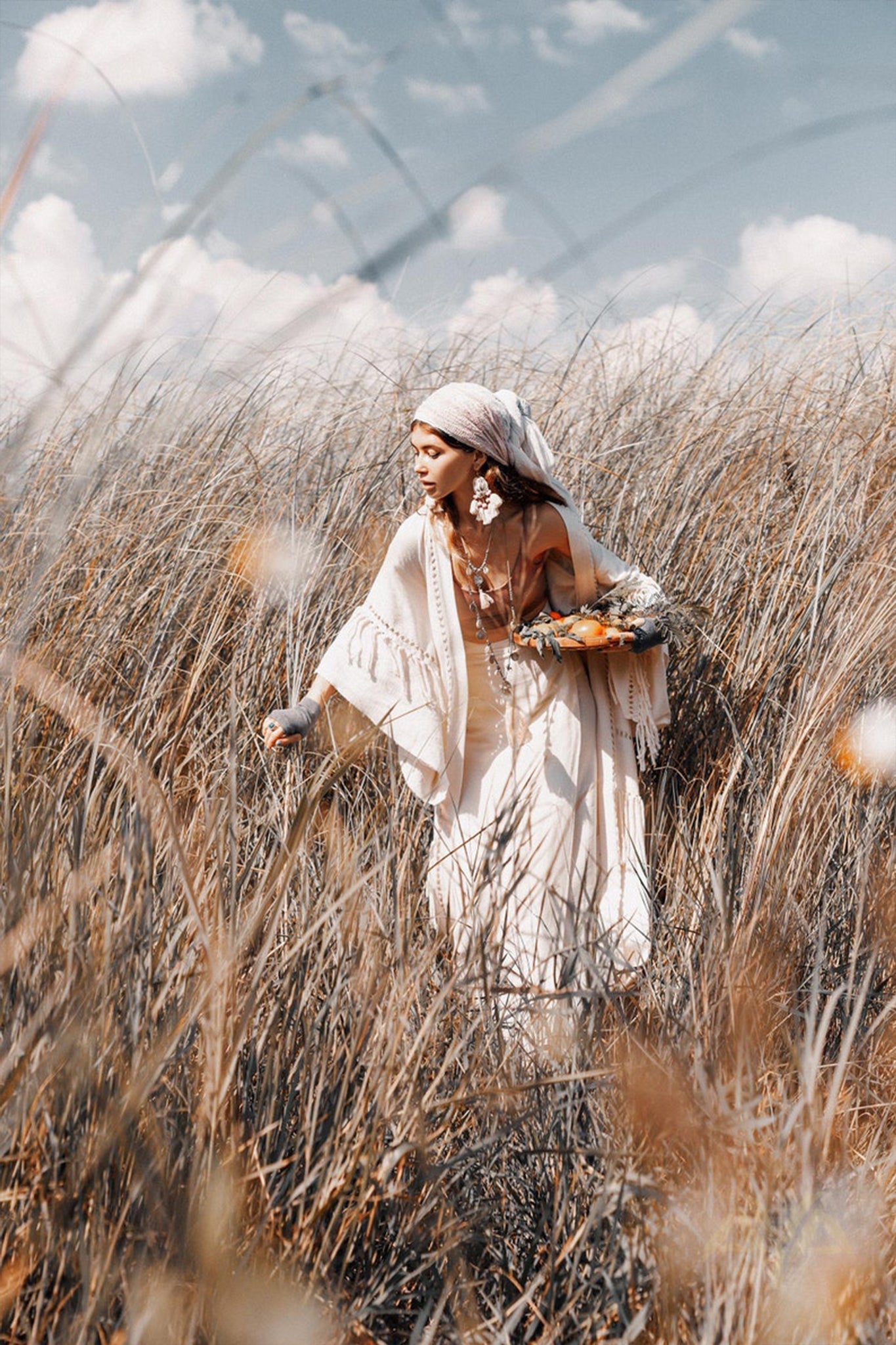 A woman dressed in a white dress and shawl gracefully walks through a field of tall grass, wearing the AYA Sacred Wear's Boho Kimono Cardigan complemented by a stylish headscarf. She gazes downward while holding a bouquet of flowers, as the sky, dotted with scattered clouds, enhances the serene atmosphere.