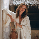 A woman with long hair in an AYA Sacred Wear Boho Kimono Cardigan stands leaning against a wall. She holds a woven jug. The background features a rustic thatched roof and wooden elements, creating an earthy, natural atmosphere.