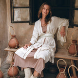 A woman in a Boho Kimono Cardigan by AYA Sacred Wear sits on a wooden bench, surrounded by decorative pottery. The background features an open window with rustic wooden shutters. She is wearing beige boots and a light dress underneath the cardigan.