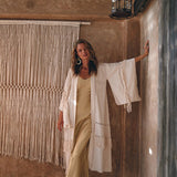 A woman stands in a rustic setting, wearing a light yellow dress and an AYA Sacred Wear Boho Kimono Cardigan. She is leaning against a textured wall with macrame decor. Above her, a lantern hangs from the wooden ceiling as natural light filters into the scene.