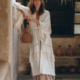 In a rustic setting over a wooden floor, a woman stands under the thatched roof wearing AYA Sacred Wear's Boho Kimono Cardigan, featuring handwoven natural cotton with fringe detailing. Holding a large jug amidst similar jugs, her light brown hair and white boots perfectly complement her look.