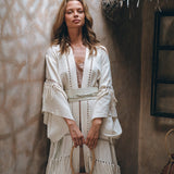 A woman in a white fringed dress stands against a textured wall, draped in AYA Sacred Wear's Boho Kimono Cardigan. Holding a rustic clay pot, she is surrounded by earthy tones and a thatched roof, creating a natural and serene atmosphere.