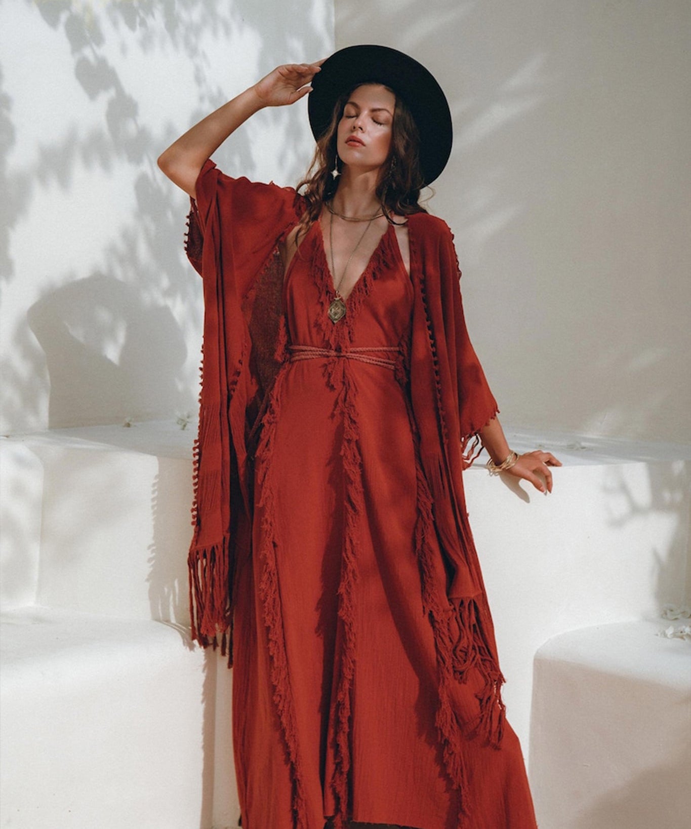 A person in a long, AYA Sacred Wear Boho Maxi Dress, also known as the Red Organic Goddess Dress or Cotton Bridesmaid Dress, complemented by a wide-brimmed hat stands against a light-colored wall. They are adorned with layered necklaces and bracelets, with sunlight casting soft shadows around them.