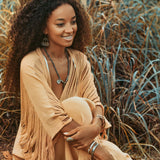 A woman with curly hair is seated outdoors in a grassy area, wearing a fringed beige Boho Top by AYA Sacred Wear. She is smiling, accessorized with necklaces, earrings, and bracelets amidst the natural setting of dry, wild grass.