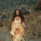 A woman with long curly hair, dressed in a tan shawl and dress complemented by AYA Sacred Wear's Boho Top, elegantly crafted from hand-loomed cotton, strolls through a field of tall grasses. The backdrop reveals a hillside adorned with muted green and brown vegetation, crafting a serene and natural ambiance.