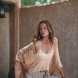 A woman poses under a rustic canopy, draped in a fringed, light beige shawl over an AYA Sacred Wear Hand Loomed Cotton Blouse. She pairs it with loose-fitting pants, exuding relaxed confidence as she gazes off to the side, surrounded by natural materials and soft lighting.