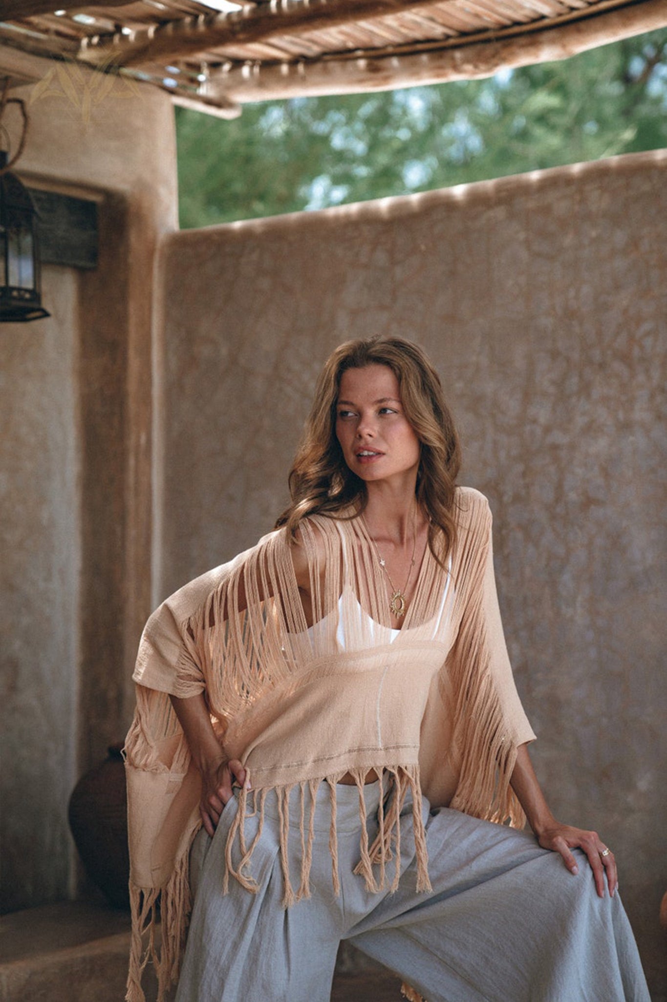 A woman poses under a rustic canopy, draped in a fringed, light beige shawl over an AYA Sacred Wear Hand Loomed Cotton Blouse. She pairs it with loose-fitting pants, exuding relaxed confidence as she gazes off to the side, surrounded by natural materials and soft lighting.