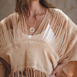 A woman is wearing a fringed beige shawl over an AYA Sacred Wear Boho Top, accessorized with a sun-shaped pendant necklace. Her long, wavy hair cascades down her shoulders as she stands against a neutral, blurred background.