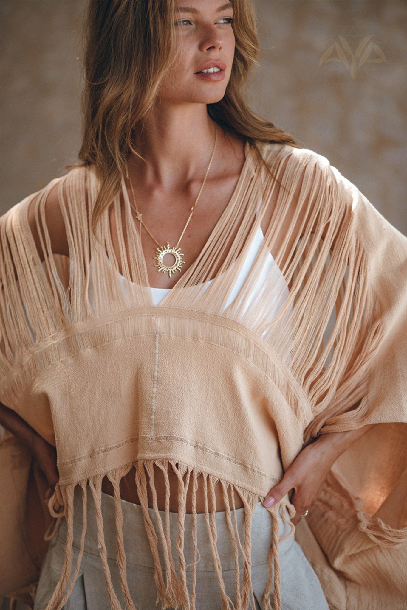 A woman is wearing a fringed beige shawl over an AYA Sacred Wear Boho Top, accessorized with a sun-shaped pendant necklace. Her long, wavy hair cascades down her shoulders as she stands against a neutral, blurred background.