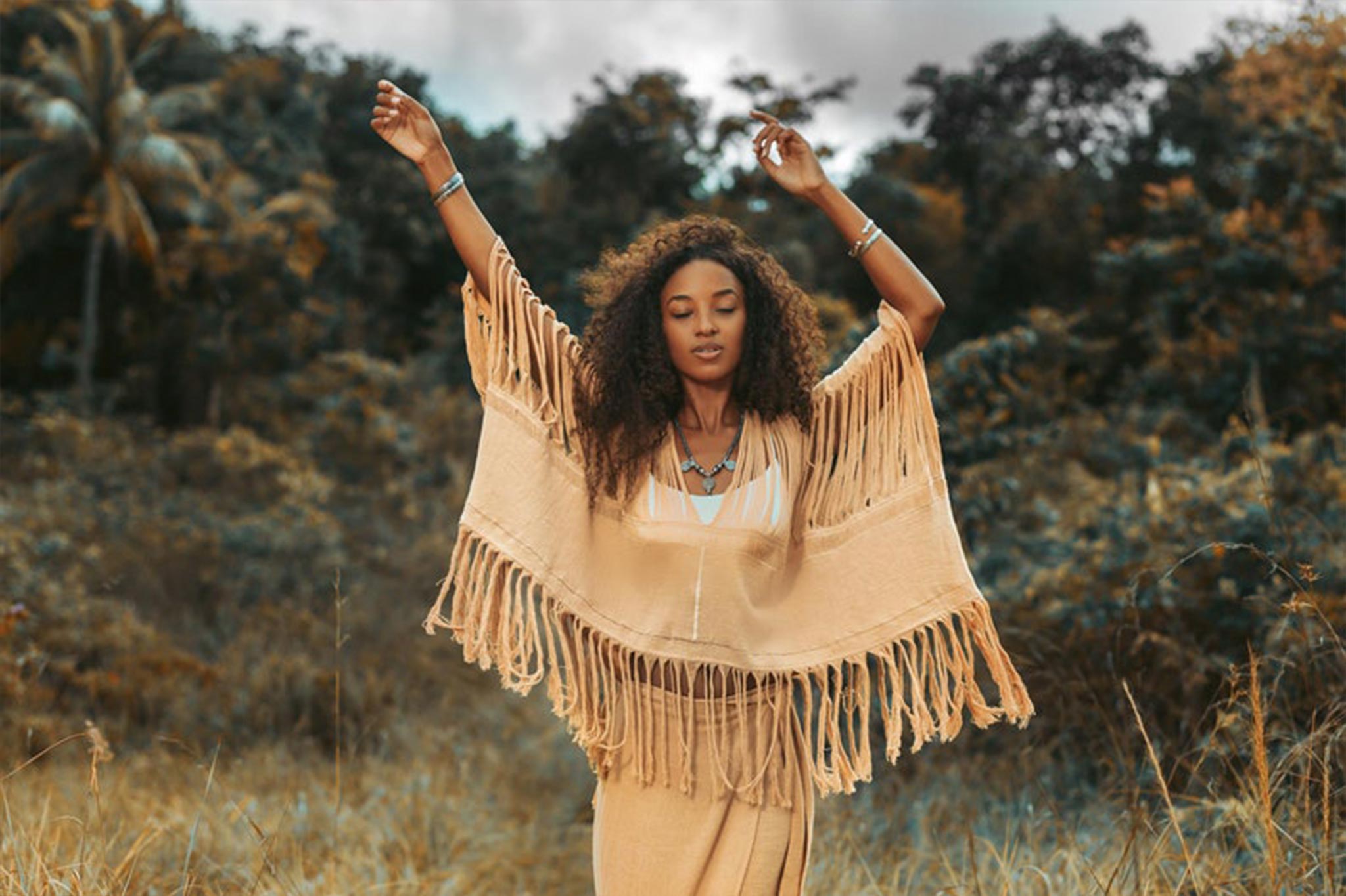 Wearing a Boho Top from AYA Sacred Wear, a woman with curly hair stands in a field, effortlessly draped in the hand-loomed cotton blouse. With her arms raised and surrounded by trees beneath an overcast sky, she embodies a serene and natural atmosphere.