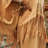 A person wearing the AYA Sacred Wear Boho Top, a beige hand-loomed cotton blouse with bohemian fringe details, stands with hands on hips. A silver bracelet adorns their wrist. Dry grass in the background enhances the warm, earthy ambiance that evokes sustainable elegance.
