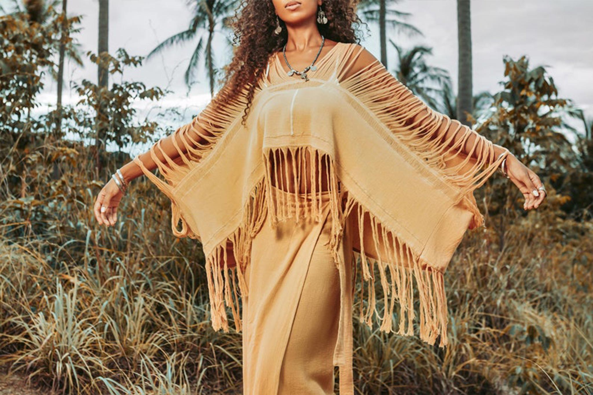 A person stands outdoors amid lush greenery and palm trees, wearing a tan fringed piece from AYA Sacred Wear—a hand-loomed cotton blouse known as the Boho Top. Paired with a matching long skirt, this ensemble fits perfectly into the bohemian-inspired scene set against an overcast sky.