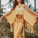 A person stands outdoors amid lush greenery and palm trees, wearing a tan fringed piece from AYA Sacred Wear—a hand-loomed cotton blouse known as the Boho Top. Paired with a matching long skirt, this ensemble fits perfectly into the bohemian-inspired scene set against an overcast sky.