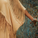A person wearing the AYA Sacred Wear Boho Top, a light brown hand-loomed cotton blouse, stands in a natural setting, gently touching the leaves of a plant. Their arm is adorned with silver bracelets, and the scene evokes a sense of tranquility and connection with nature. The hand-loomed cotton adds an earthy, sustainable touch.