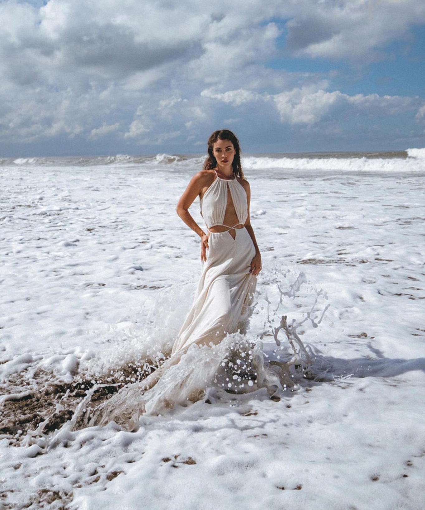 A woman in a Boho Wedding Dress for Women, an open-back sides belly dress designed by AYA Sacred Wear, stands on a beach, surrounded by foamy waves. The sky above is cloudy, and the ocean creates a dramatic backdrop.