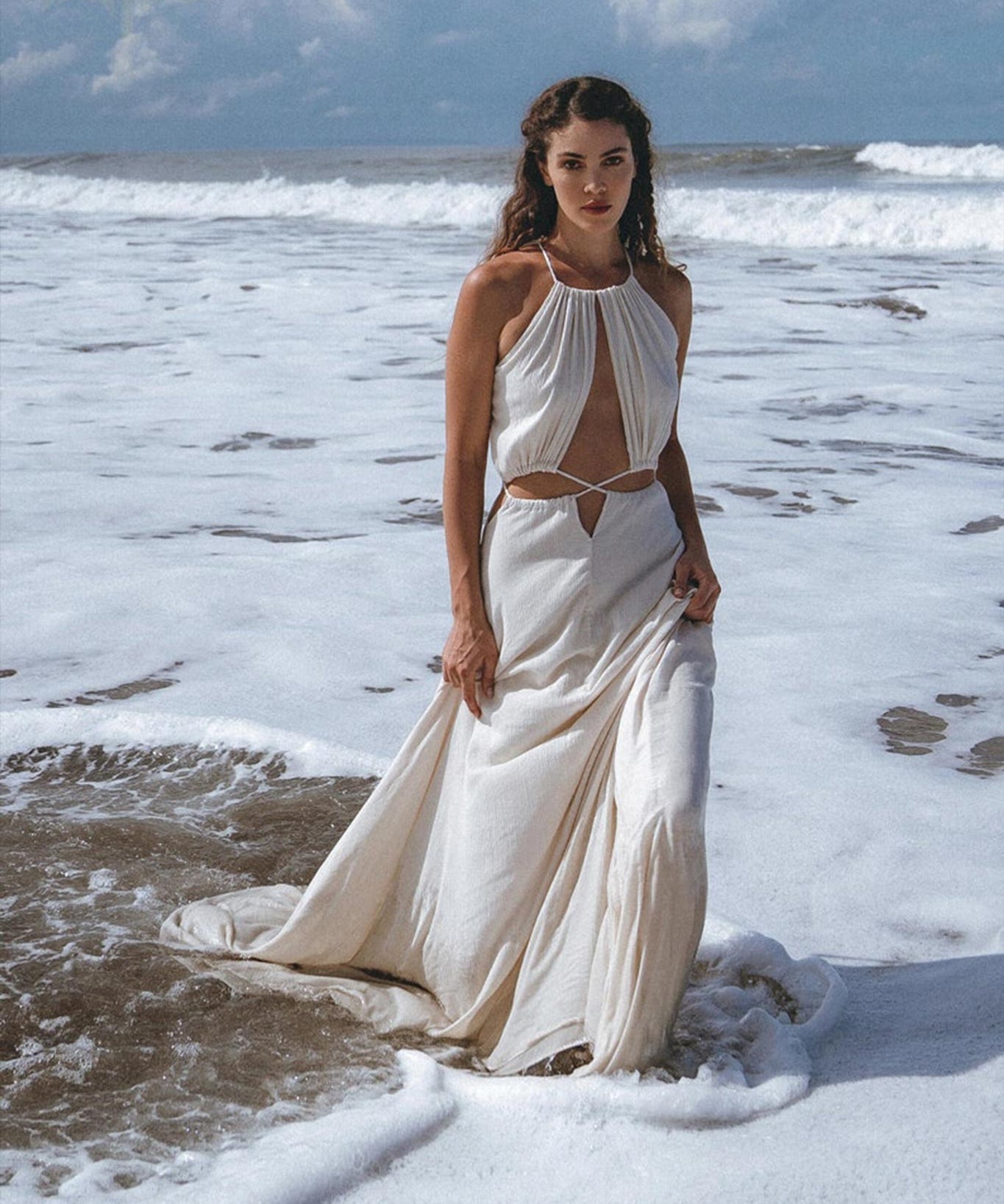 A woman stands gracefully in shallow ocean water, adorned in a Boho Wedding Dress from AYA Sacred Wear, featuring an open back and sides belly design with handwoven fabric. The sky is partly cloudy, and waves gently crash around her feet in a serene coastal setting reminiscent of Ayurvedic harmony with nature.