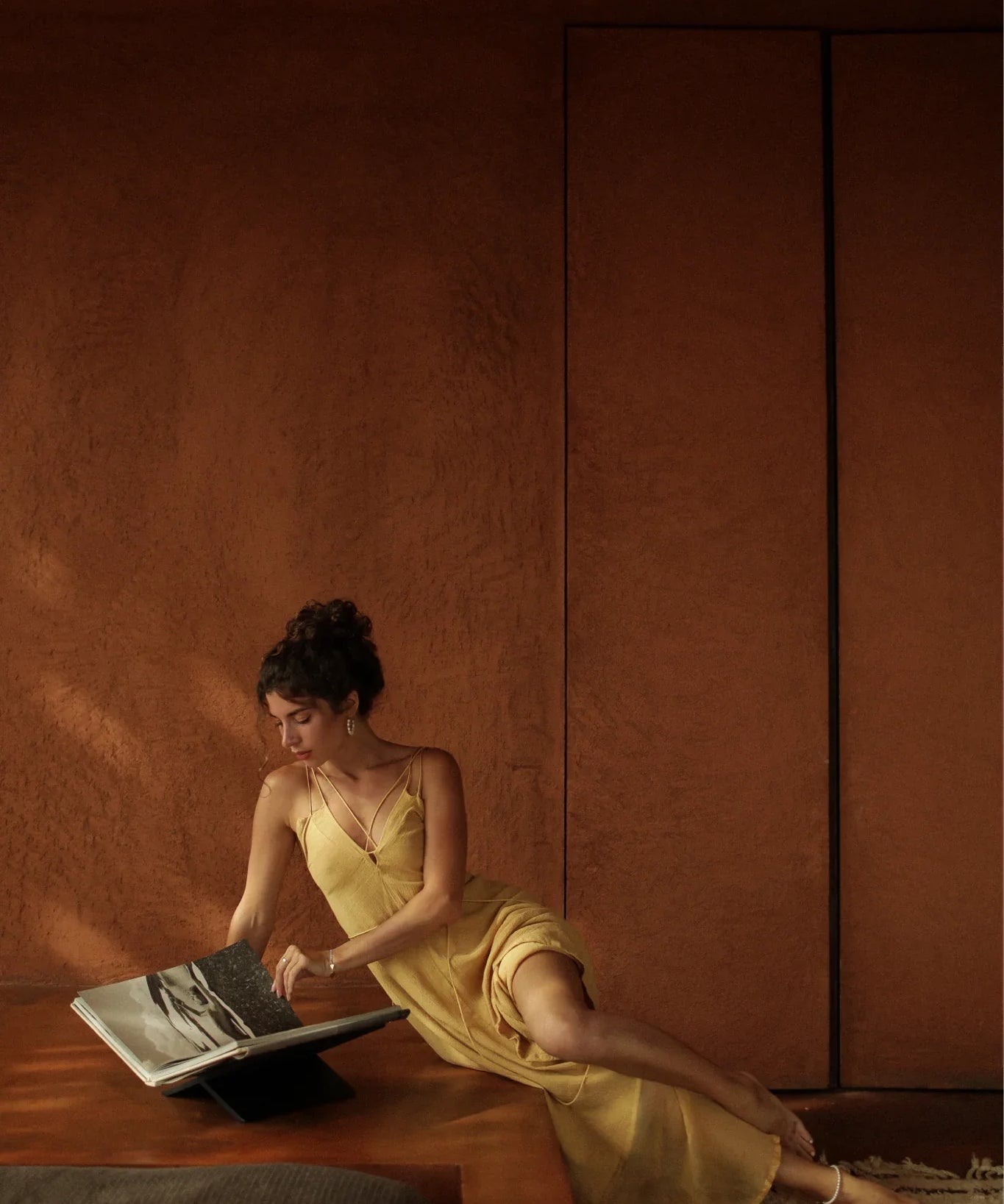 A woman in the Powder Yellow Padma Dress by AYA Sacred Wear sits on a patterned rug, leaning against wood, reading a large book in a warmly lit room with earthy brown walls.