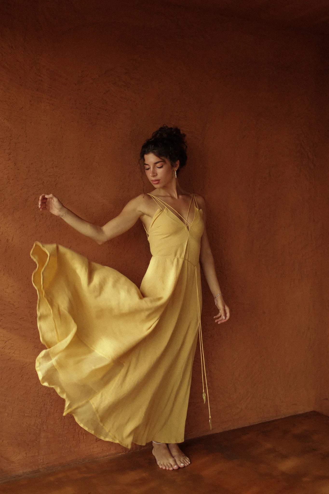 A woman in the Powder Yellow Padma Dress by AYA Sacred Wear stands barefoot against a textured brown wall, gently lifting the hem of her sleeveless boho backless dress and gazing downward with a serene expression.