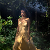 Amidst lush greenery and soft sunlight, a woman in AYA Sacred Wear's Powder Yellow Padma Dress—a boho backless, sleeveless elegant dress—stands with a serene and contemplative presence, gazing forward.