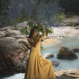 A woman stands on rocks near a serene stream, wearing the Powder Yellow Padma Dress by AYA Sacred Wear. Surrounded by greenery and soft sunlight, she gently holds leafy branches above, creating a tranquil scene in this boho backless, sleeveless elegant dress.