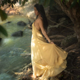 A woman in an AYA Sacred Wear Powder Yellow Padma Dress stands by a stream, surrounded by lush greenery. As she holds the hem of the boho backless and sleeveless elegant dress, sunlight filters through leaves, casting gentle shadows while she gazes contemplatively at the water.