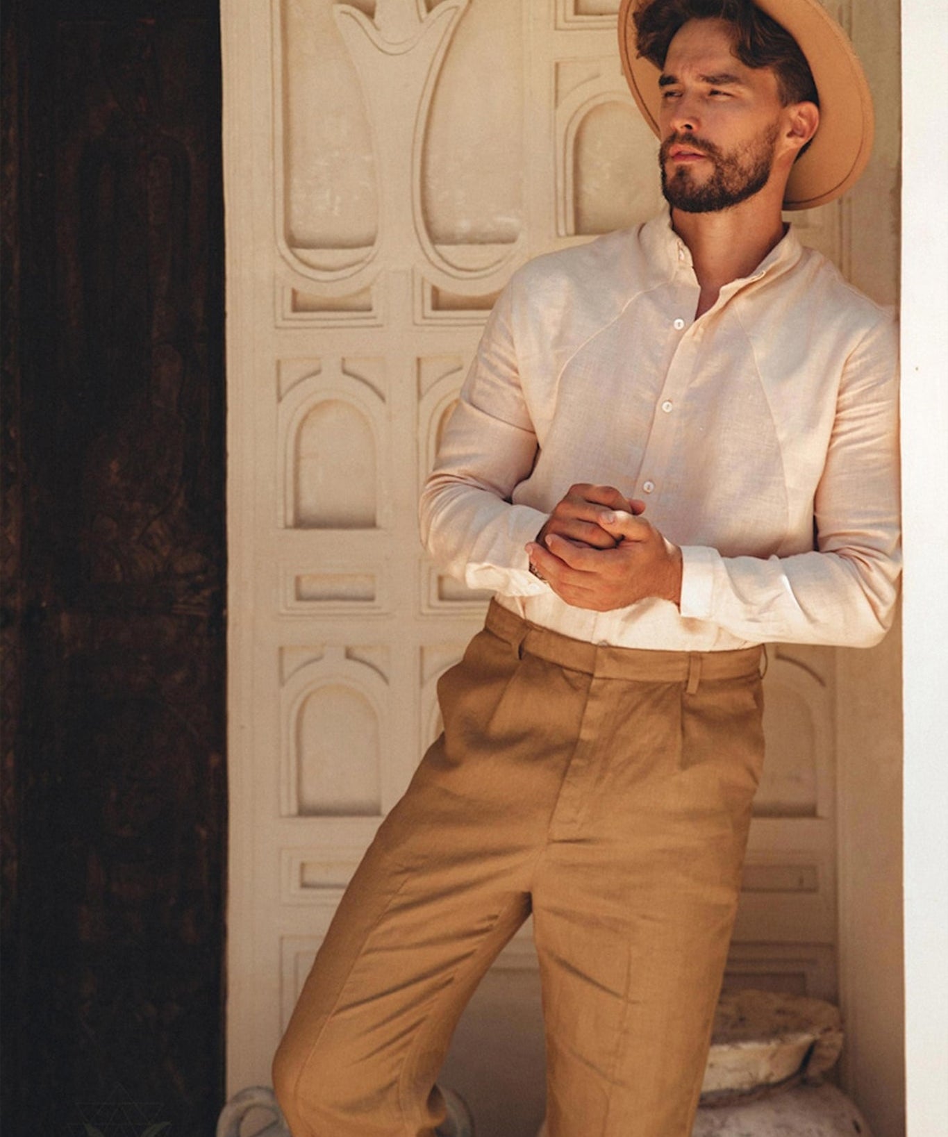 A man in a Pale Pink Linen Classic Minimalist Shirt from AYA Sacred Wear and tan trousers leans against a wall with decorative patterns. He has a contemplative expression beneath his wide-brimmed hat. Large pots adorn the background, complementing the natural charm of his botanically dyed attire.