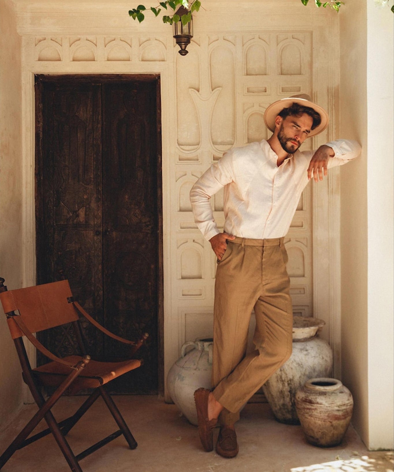 Dressed in a Pale Pink Linen Classic Minimalist Shirt from AYA Sacred Wear and tan pants, a man complements his outfit with a beige hat as he leans against an ornate rustic wall. Large clay pots surround him, while a wooden chair sits to his left, and green leaves rustle softly under the glow of a hanging lantern.