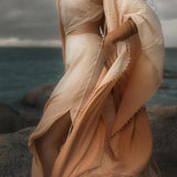 Standing on rocky sea terrain, a person wears the AYA Sacred Wear Powder Pink Boho Poncho, an elegant handmade bohemian cape with textured edges. Layered necklaces and bracelets add charm against the ocean and moody sky backdrop.