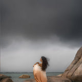 A woman in a flowing Powder Pink Boho Poncho by AYA Sacred Wear stands on rocky terrain by the sea. Stormy gray clouds loom overhead, and her long hair blows in the wind, enhancing the dramatic atmosphere.