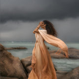 A woman stands on rocks by the sea in AYA Sacred Wear's Powder Pink Boho Poncho, a flowing ombre sustainable mantel. Her hair blows as she gazes at the stormy sky, with dark clouds and a turbulent ocean in the background.