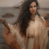A woman with curly hair stands by the sea in a Powder Pink Boho Poncho from AYA Sacred Wear. The wind gently stirs her shawl. Calm waters and rocks lie under a muted sky, reflecting the ombre tones of this sustainable wrap achieved through botanical dyeing.